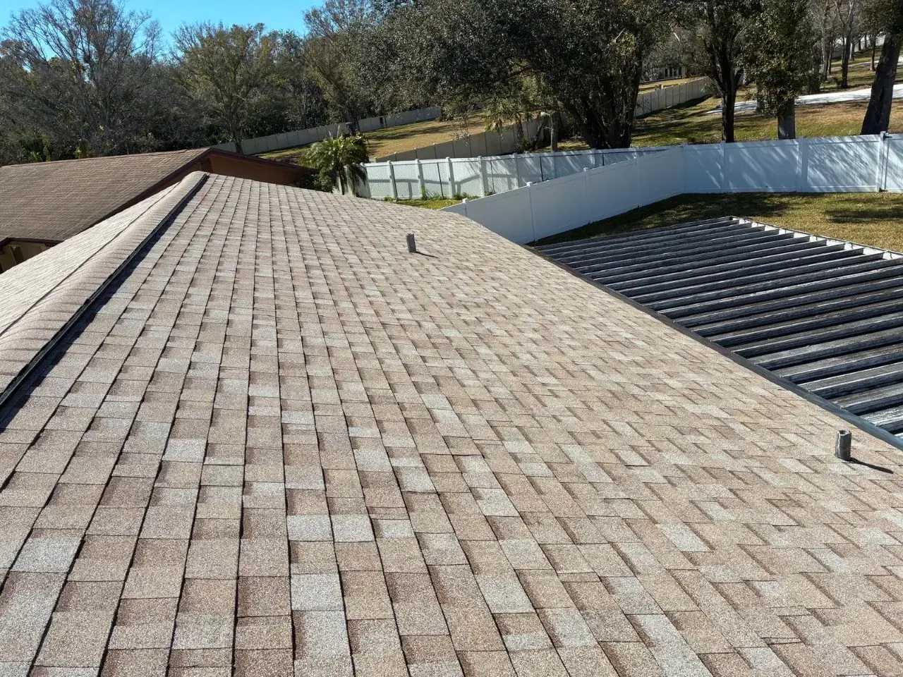 Contact Us Today Tan asphalt shingle roof beside a metal patio cover, with a white fence and trees in the background