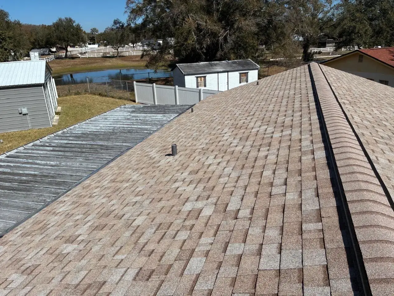 Contact Us Today Aerial view of a residential shingle roof with a ridge vent, sheds and a pond in the background
