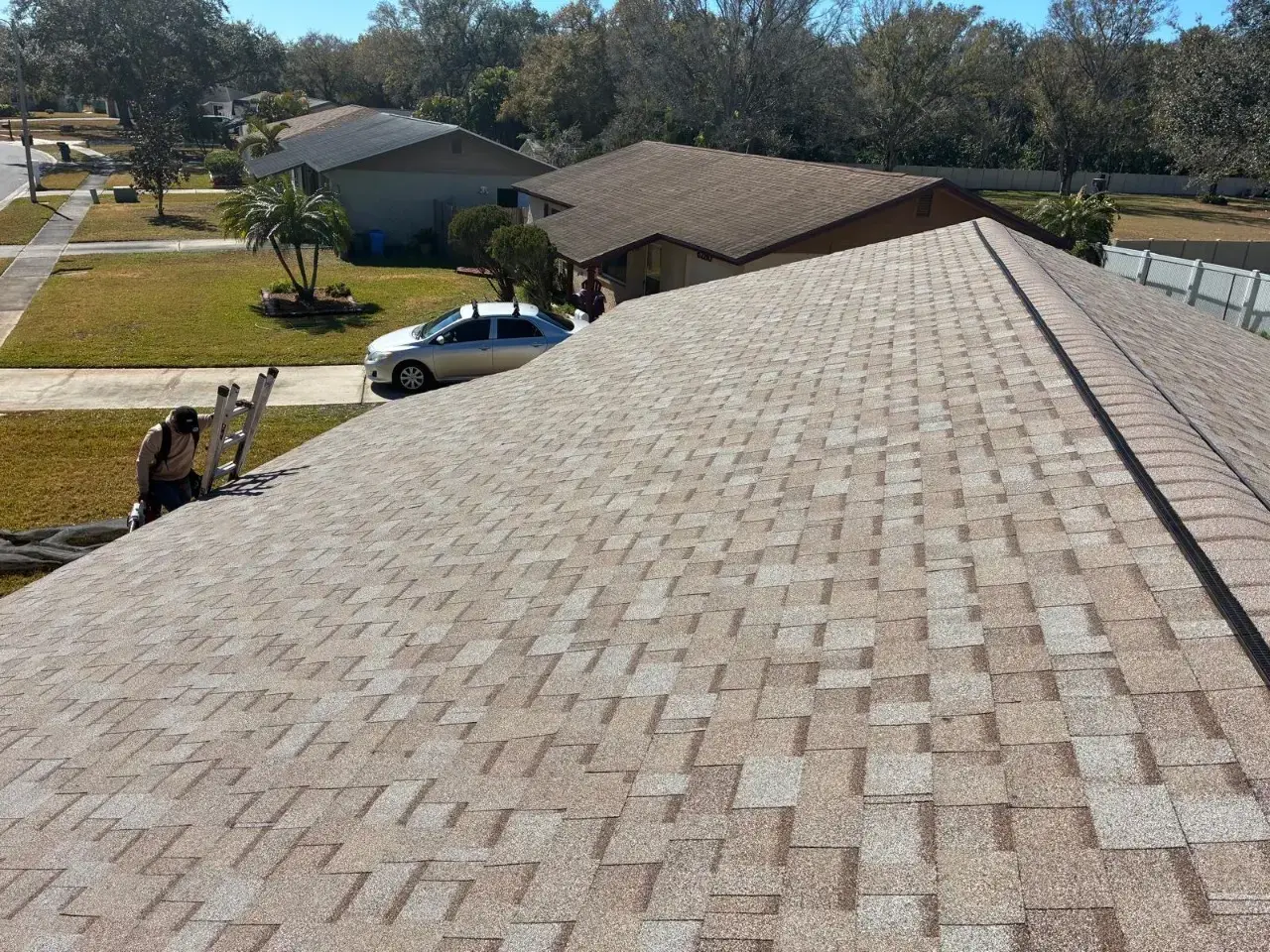 Contact Us Today Roofer stands beside a ladder at the edge of a shingled home roof in a suburban neighborhood